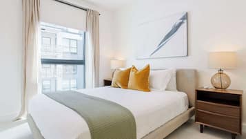 A hotel room with a large bed, two bedside tables, a window with curtains, a wall-mounted lamp, and a painting.