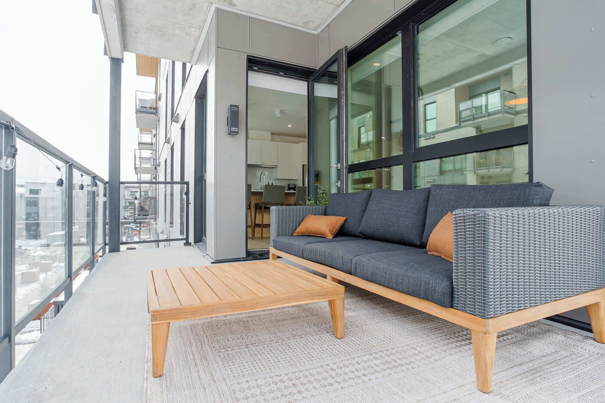 A balcony with a sofa and a wooden table, large windows, and a view of the city.