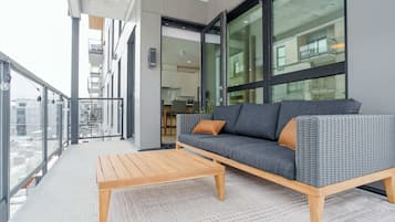 A balcony with a sofa and a wooden table, large windows, and a view of the city.