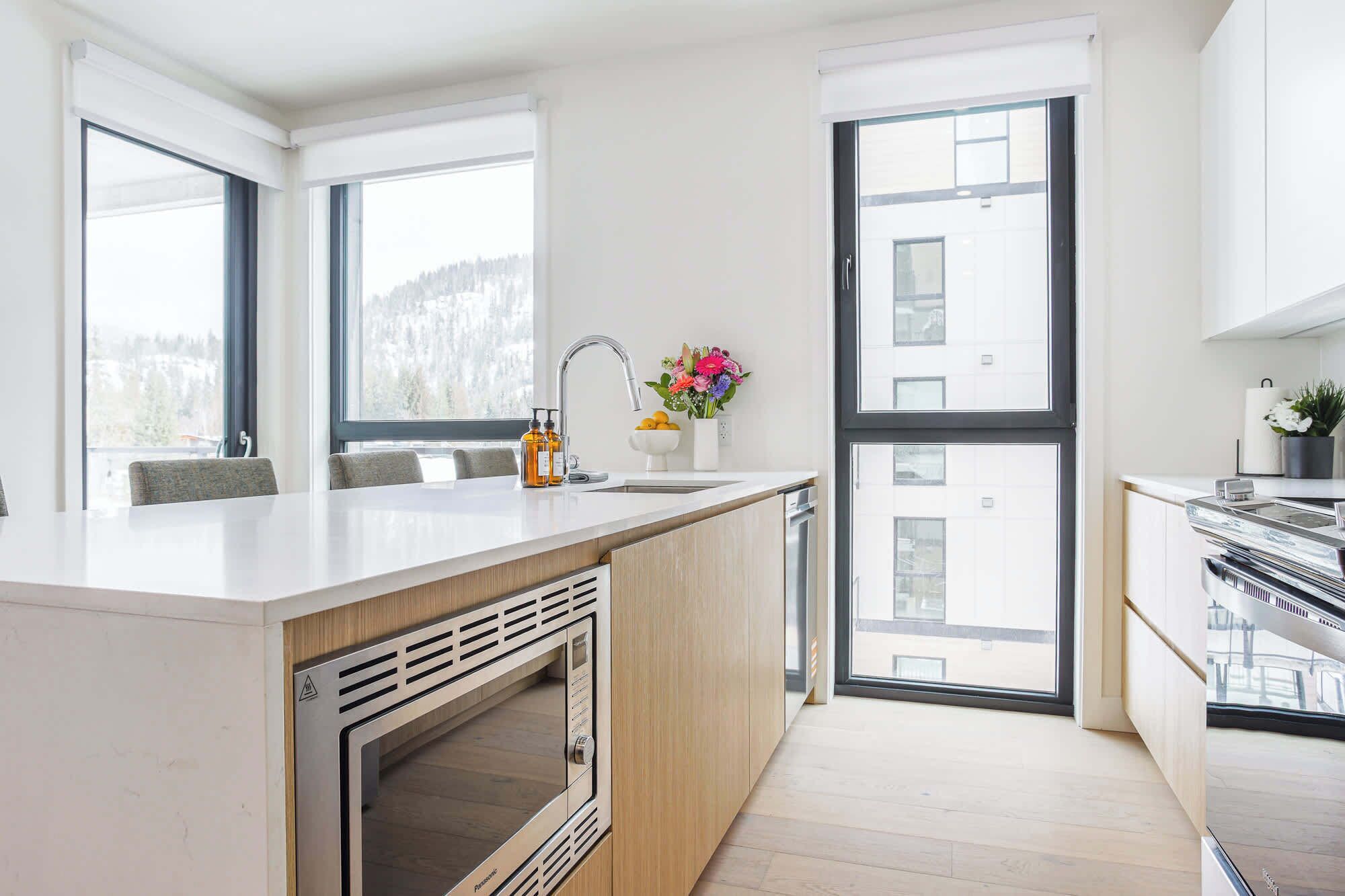 A modern kitchen with a central island, a built-in oven, and large windows offering views of the outdoors.