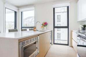 Et moderne køkken med en central ø, indbygget ovn og store vinduer med udsigt til det fri.