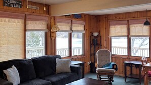 A cozy cabin interior with a dark sofa, a wooden coffee table, a chair, a ceiling fan, and large windows with wooden shutters.