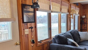 A cozy cabin interior with a wooden wall, a gray sofa, a small table, and a window with a view of a wooden deck.