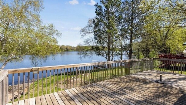 A wooden deck with a railing overlooking a calm lake, surrounded by trees and a grassy area.