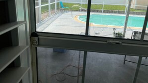 En vy genom en myggnätsdörr in mot en trädgård med en pool, en blå stol och en gräsmatta.