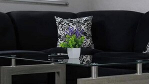 Un canapé noir agrémenté d’un coussin à motifs et d’un petit vase blanc contenant des feuilles vertes et des fleurs violettes.