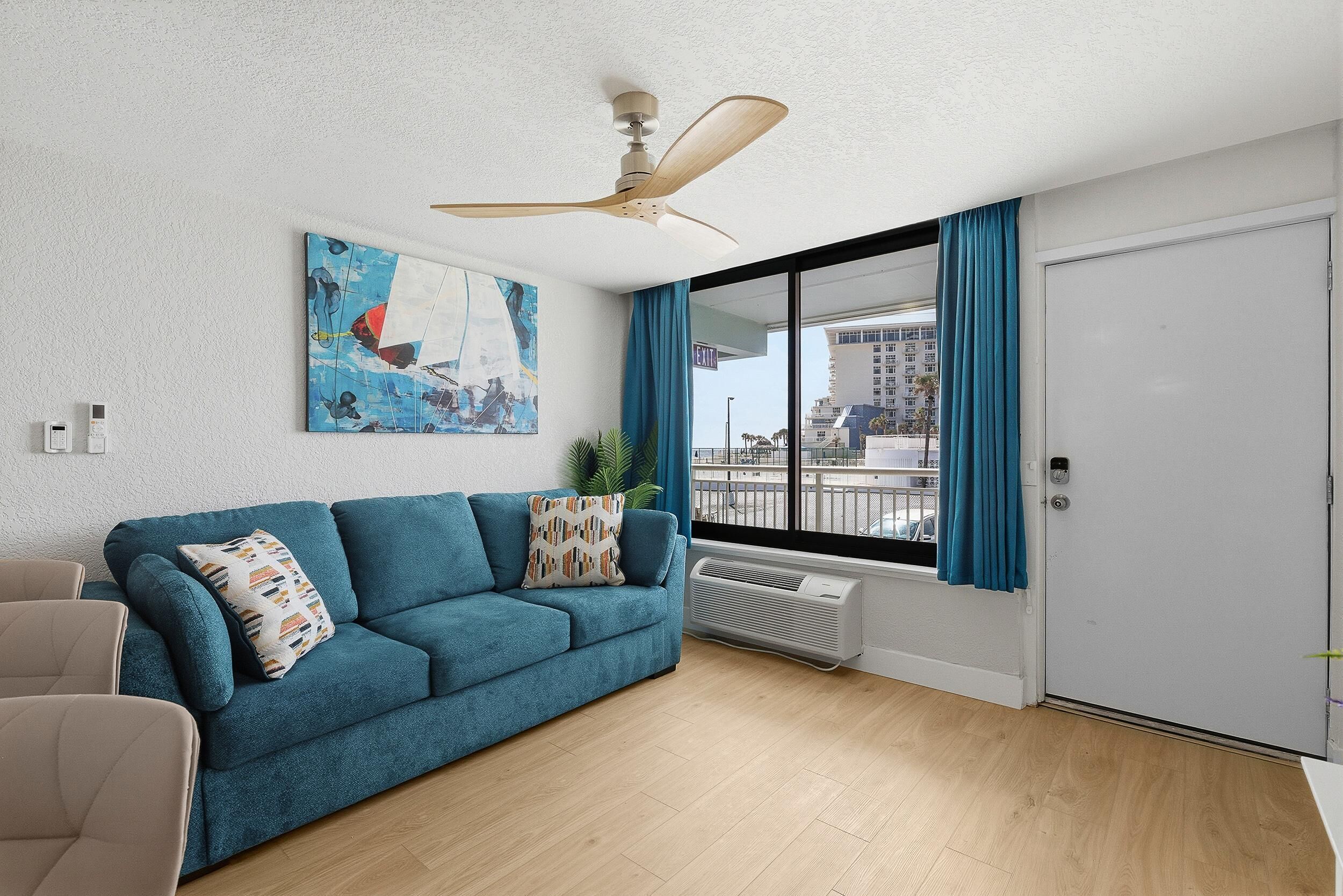 A living room with a blue sofa, a ceiling fan, a large window with curtains, and a painting on the wall.