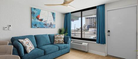 A living room with a blue sofa, a ceiling fan, a large window with curtains, and a painting on the wall.
