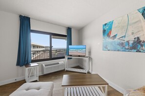 Ein Zimmer mit großem Fenster, einem Fernsehgerät auf einem weißen Schrank, einem weißen Sessel und einem Holztisch.