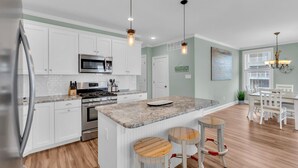 A modern kitchen with white cabinets, a central island with stools, stainless steel appliances, and a dining area with white chairs. - 10th Street Beach Block Condo (Ocean City)