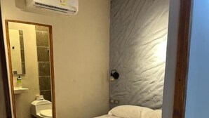 Una habitación de hotel con cama, un baño con lavamanos y espejo, y un equipo de aire acondicionado.