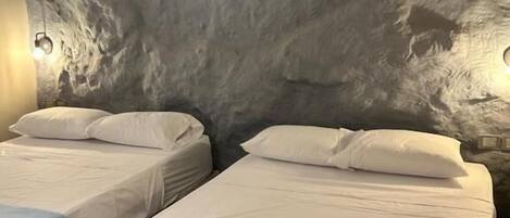 A room with two beds, each with white bedding and a blue accent. The walls have a textured, gray appearance.