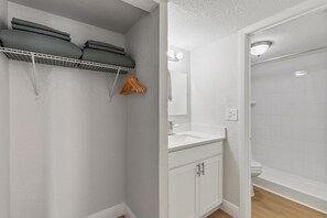 A bathroom with a white sink, mirror, and toilet, a shower area, and a storage shelf with folded towels. - Ocean Breeze Resort 109 (Daytona Beach Shores)