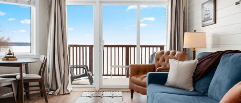 A living room with a blue sofa, a leather armchair, a wooden coffee table, and a view through a sliding glass door to a balcony.
