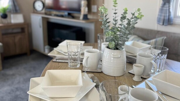 Une table à manger dressée avec de l’assiette, des bols et des verres blancs, une plante en pot et un téléviseur en arrière-plan.