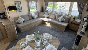 Un salon chaleureux avec un canapé d’angle, une table basse ronde aménagée pour les repas et de grandes fenêtres dotées de rideaux.