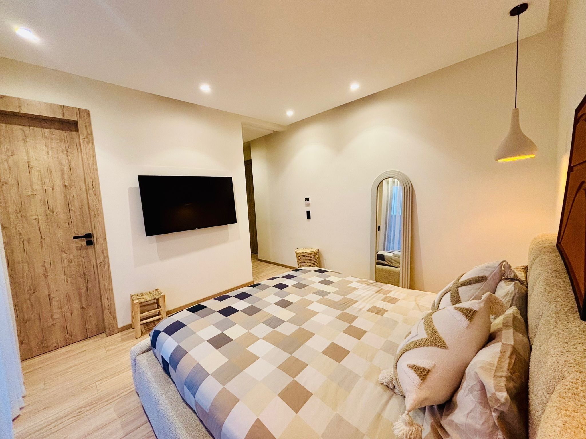 Een slaapkamer met een geruite sprei, een flatscreen televisie aan de muur, een houten deur en een spiegel.