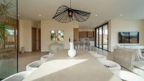 A modern dining area with a long table, white chairs, a unique pendant light, and a kitchen in the background. - Sumptuous Villa with swimming pool in the secure estate of MYSK Route d'amizmiz (Marrakech)