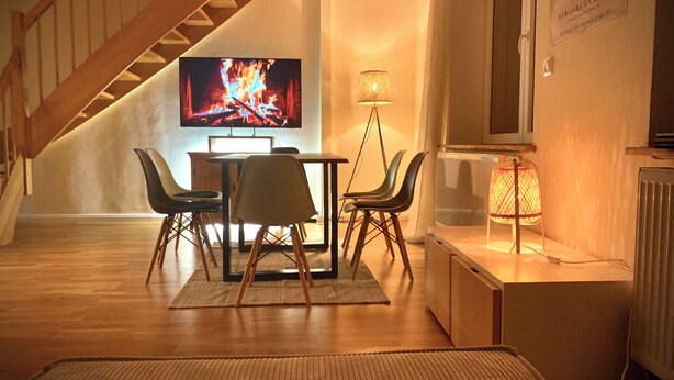 Ett vardagsrum med en trätrappa, en tv-skärm som visar en brasa, ett matbord med stolar och en golvlampa.