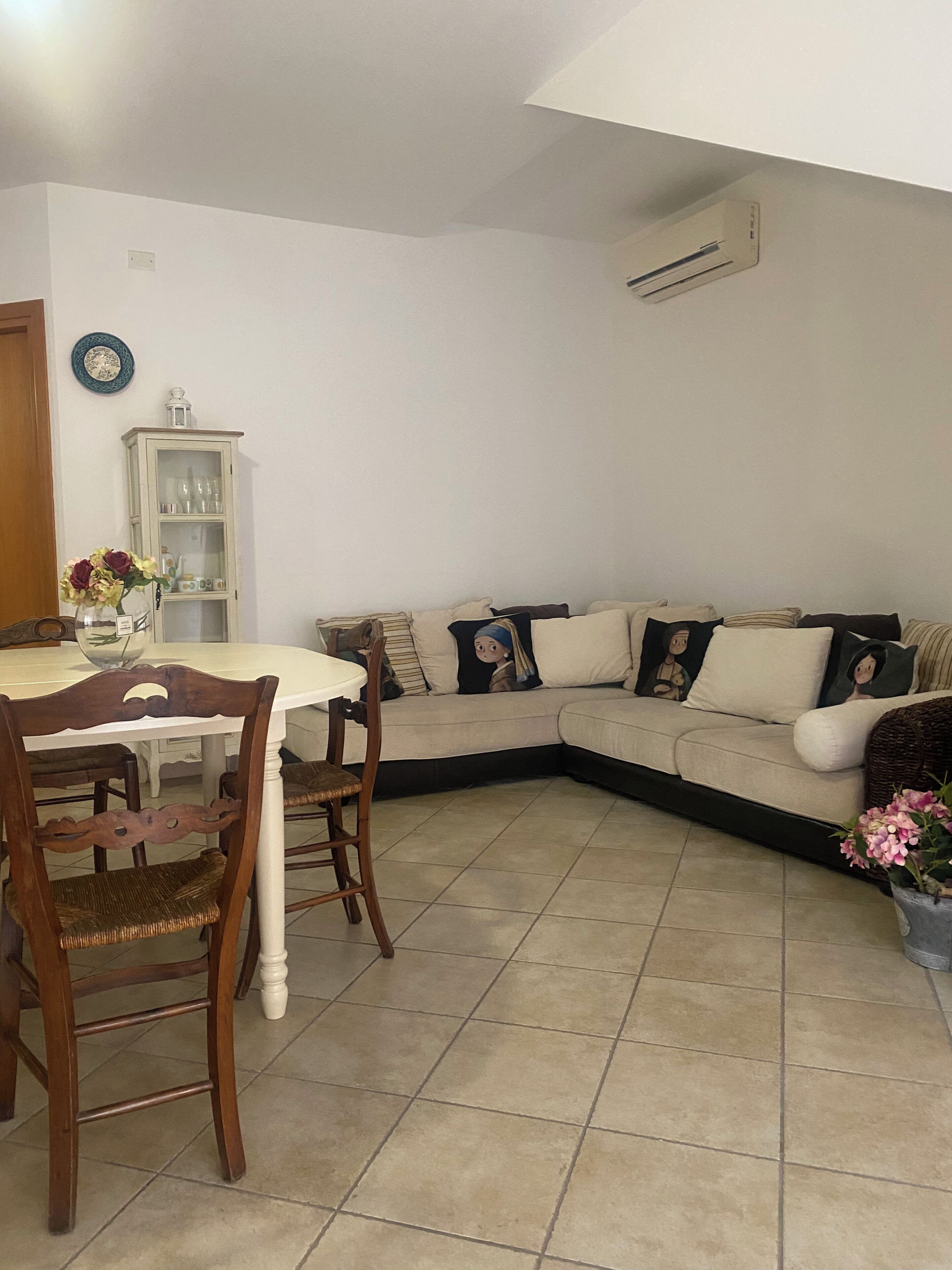 Uma sala de jantar com uma mesa redonda, cadeiras, um armário com portas de vidro, um sofá com almofadas e uma planta em vaso.