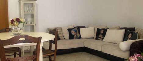 Uma sala de jantar com uma mesa redonda, cadeiras, um armário com portas de vidro, um sofá com almofadas e uma planta em vaso.