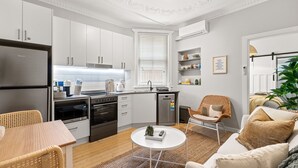 A modern kitchen with white cabinetry, a built-in oven, and a dishwasher. There is a dining area with a table and chairs, and a living area with a sofa and a coffee table. - Bondi Salt Soul - A Sepal Stay (Sydney)