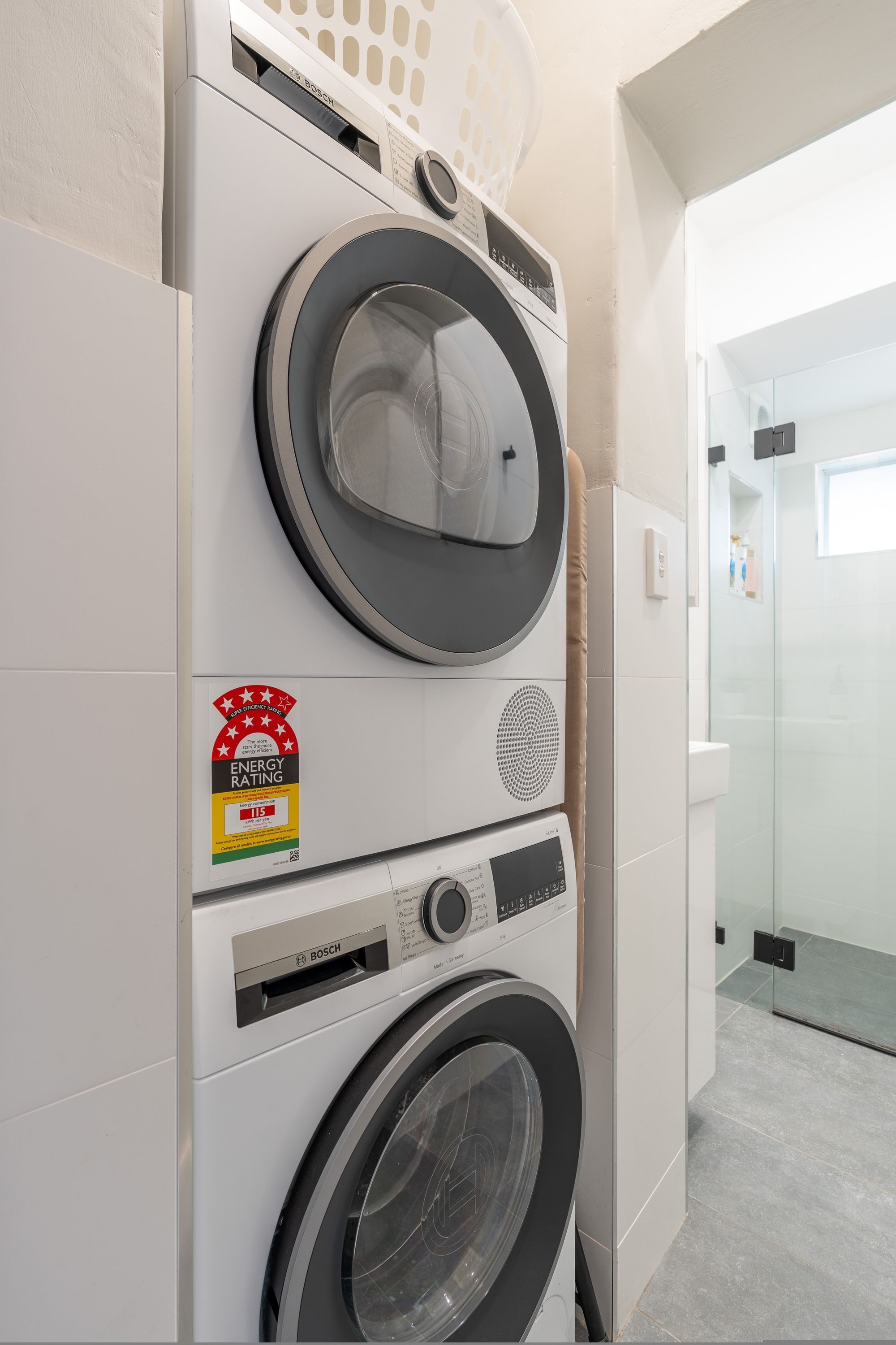A stacked washer and dryer unit with an energy efficiency rating sticker, a control panel, and a glass shower enclosure in the background.