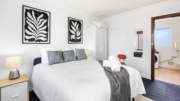 Ein ordentlich eingerichtetes Hotelzimmer mit Bett, Nachttisch, Lampe, Kunstwerk und Blick ins Badezimmer.