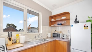 A kitchen with wooden cabinets, a microwave, a sink, and a window with a view of houses. - Skynest Rivervale - Sepal Stay (Perth)