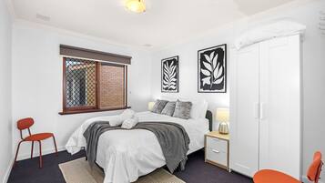 A bedroom with a bed, two chairs, a nightstand, a wardrobe, and artwork on the wall.