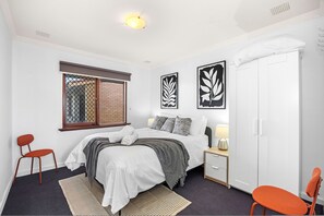 A bedroom with a bed, two chairs, a nightstand, a wardrobe, and artwork on the wall. - Skynest Rivervale - Sepal Stay (Perth)