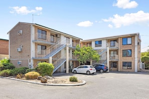 A multi-story building with a central staircase, parking lot, and a small landscaped area with rocks and shrubs. - Skynest Rivervale - Sepal Stay (Perth)