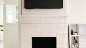 Un salon avec une cheminée en briques blanches, un téléviseur à écran plat fixé au mur et une étagère blanche ornée de plantes.