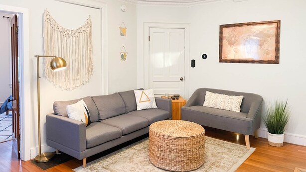 Un salon avec un canapé gris, un pouf rond en osier, une lampe sur pied, une décoration murale en macramé, un tableau représentant une carte du monde et une plante en pot.