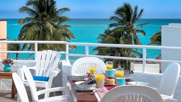 A balcony with white chairs, a table with breakfast items, and a view of the ocean and palm trees. - Casa 4 Estaciones (Puerto Morelos)