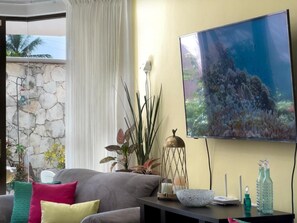 A living room with a stone wall, a grey sofa with colorful cushions, a TV on the wall, and a decorative lamp. - Casa 4 Estaciones (Puerto Morelos)
