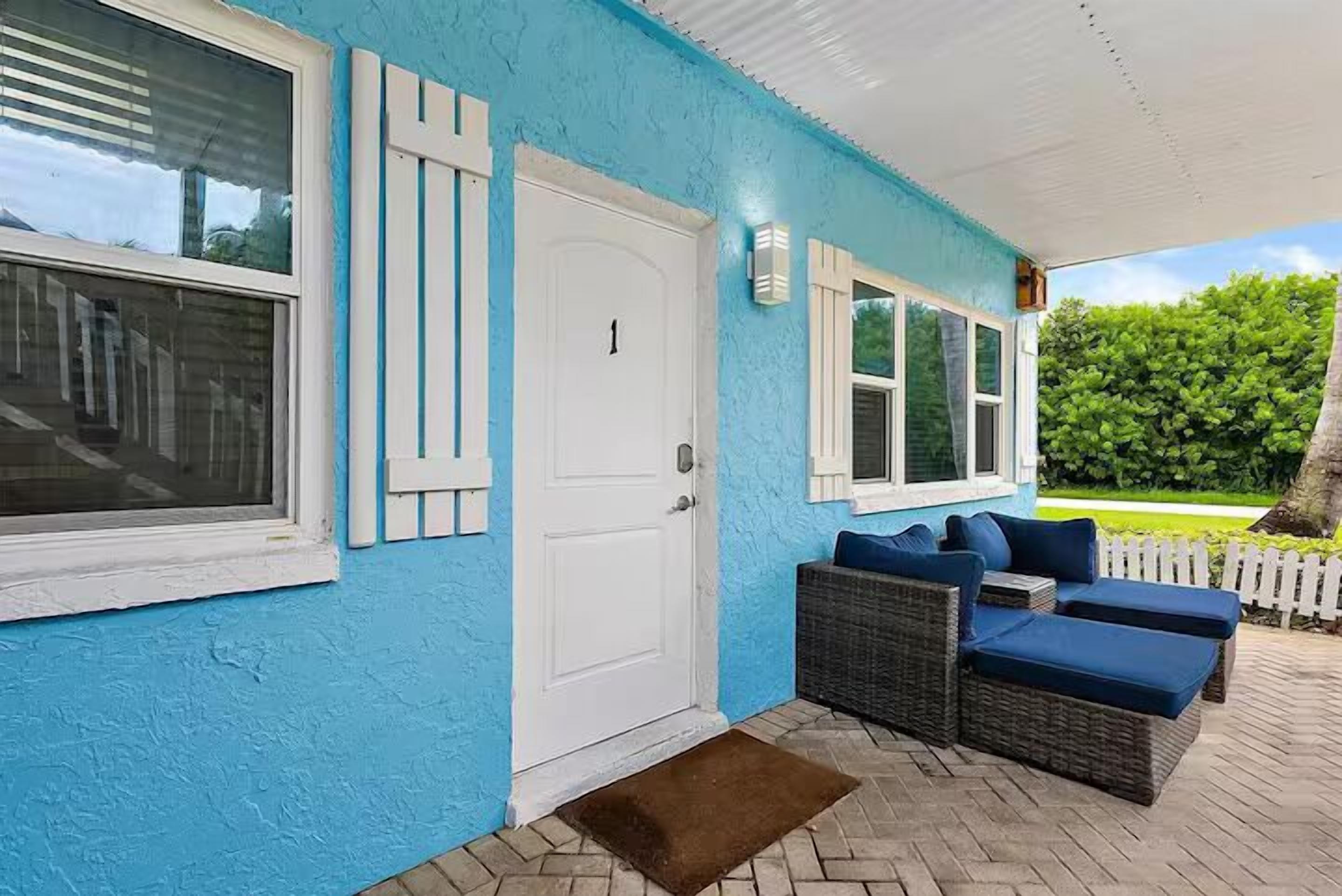 A covered porch with a blue wall, white shutters, a white door, and wicker furniture with blue cushions.