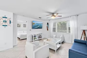 A modern living room with a ceiling fan, a TV, a white sofa, a blue sofa, and a bed in the background. - Hutchinson Island Beach Retreat | Pets Welcome (Fort Pierce)