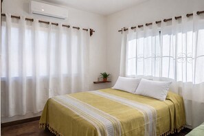 Un dormitorio con cama, cortinas, una planta en maceta y un equipo de aire acondicionado.