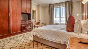 A hotel room with a large bed, wooden wardrobe, a desk with a chair, a television, and a window with curtains. - Deluxe One-Bedroom Apartment (Sandton)