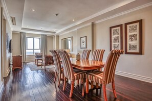 A spacious dining area with a wooden table and striped chairs, a large window with curtains, and framed artwork on the walls. - Deluxe One-Bedroom Apartment (Sandton)