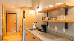 A modern kitchen with wooden cabinets, a stainless steel refrigerator, a coffee maker, and a shelf with dishes and jars.