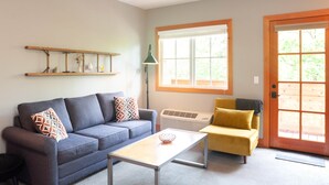 A living room with a grey sofa, a yellow armchair, a coffee table, a window with blinds, and a wooden door. - Studio 2 @ The Suites on Main (Leavenworth)