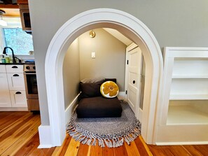 Een gezellige kamer met een klein bed, een decoratief vloerkleed, en een keuken zichtbaar door een boog.