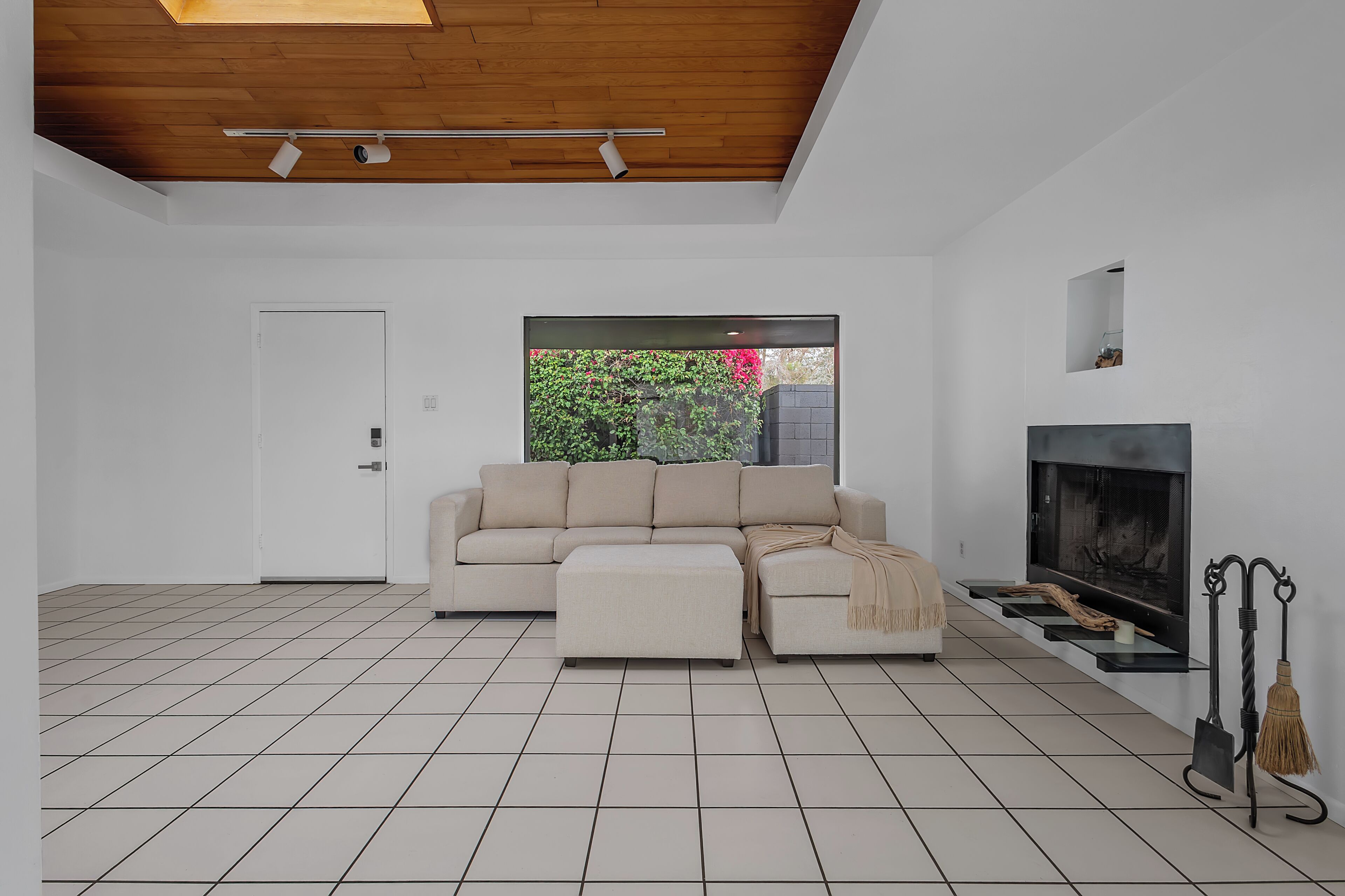 A modern living room with a sofa, ottoman, fireplace, and a view of greenery outside.