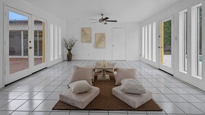 Una habitación amplia con piso de baldosas, ventilador de techo, mesa de comedor con sillas y ventanas grandes que funcionan como puertas.