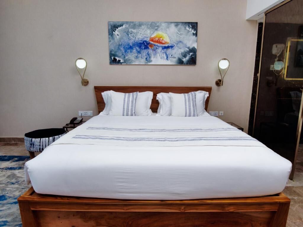 A neatly made bed with white bedding and a wooden headboard, a painting above the bed, and two wall-mounted lamps on either side.