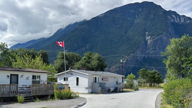 A road leading to a small building with a flag, surrounded by greenery and a mountain in the background. - Retasket Lodge & RV Park (Lillooet)