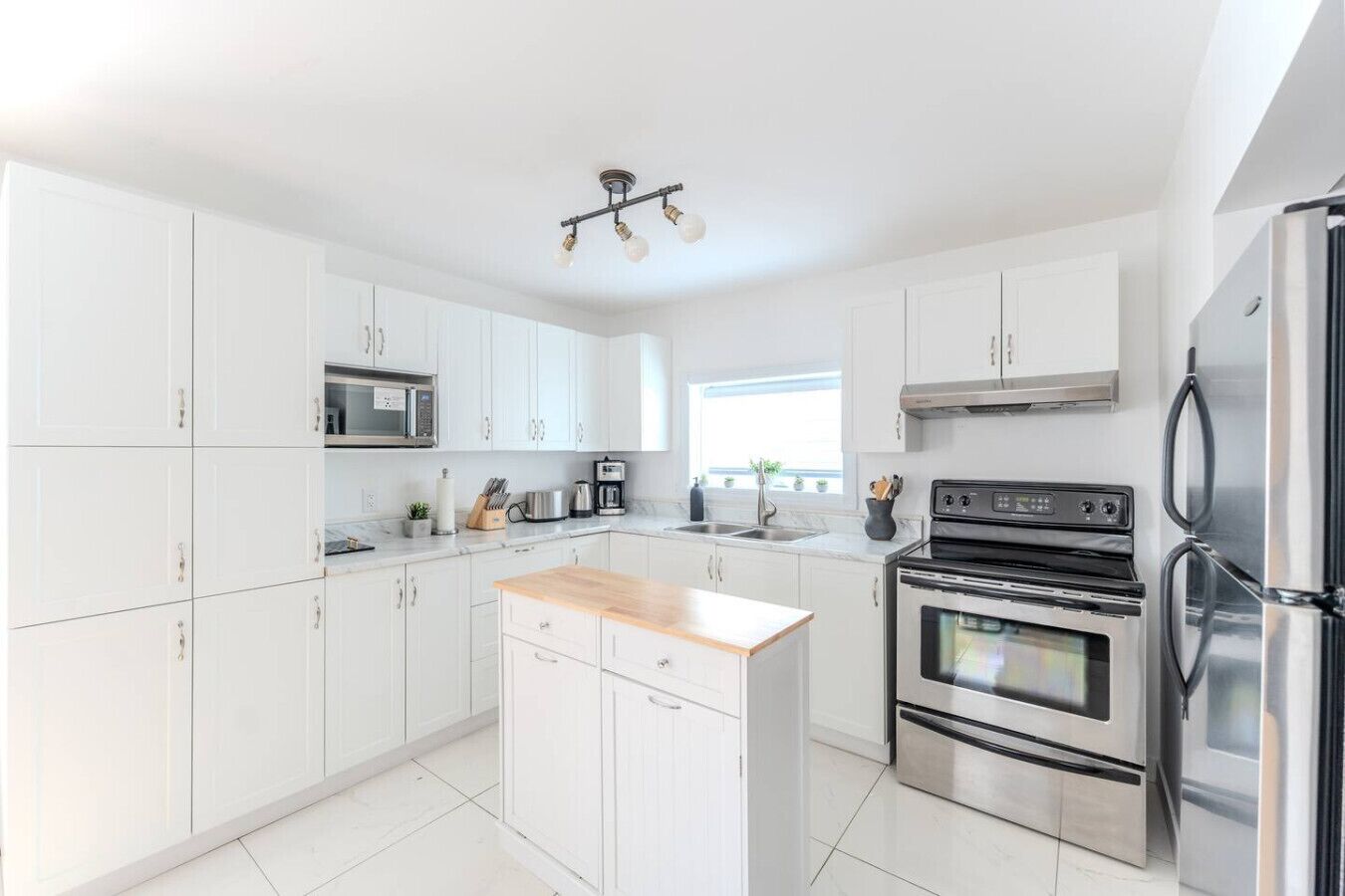 A modern kitchen with white cabinets, stainless steel appliances, a wooden island, and a window providing natural light.