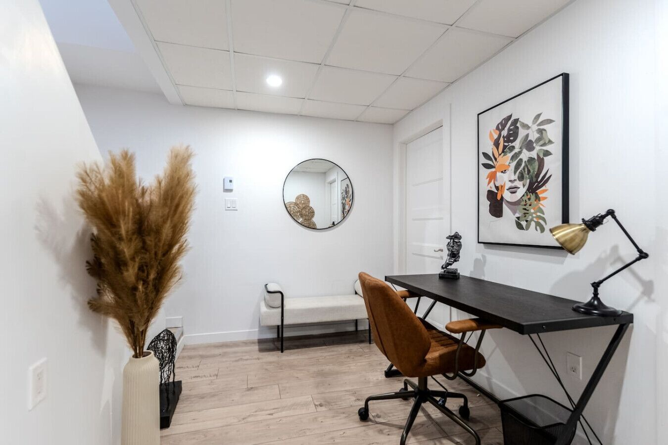 Ein Zimmer mit Schreibtisch, Stuhl, Spiegel, Kunstwerken und einer Vase mit Trockenpflanzen.